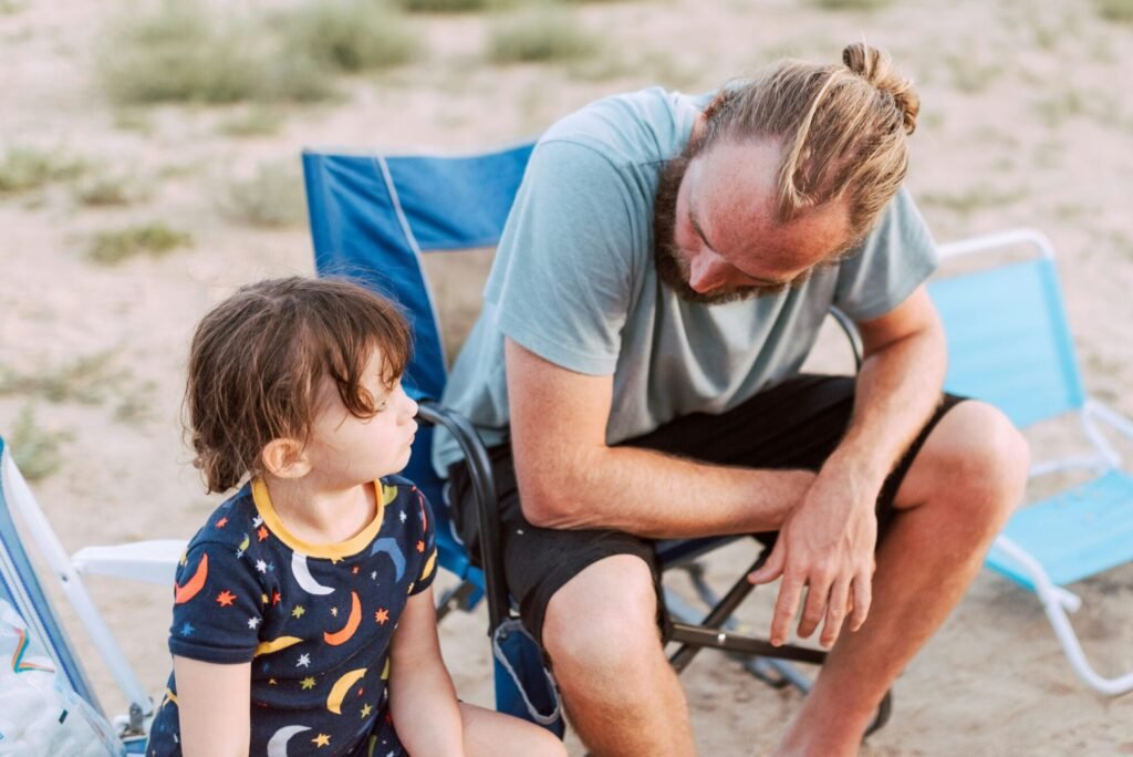 A father demonstrates positive parenting solutions by calmly talking to his upset daughter while sitting on the beach.
